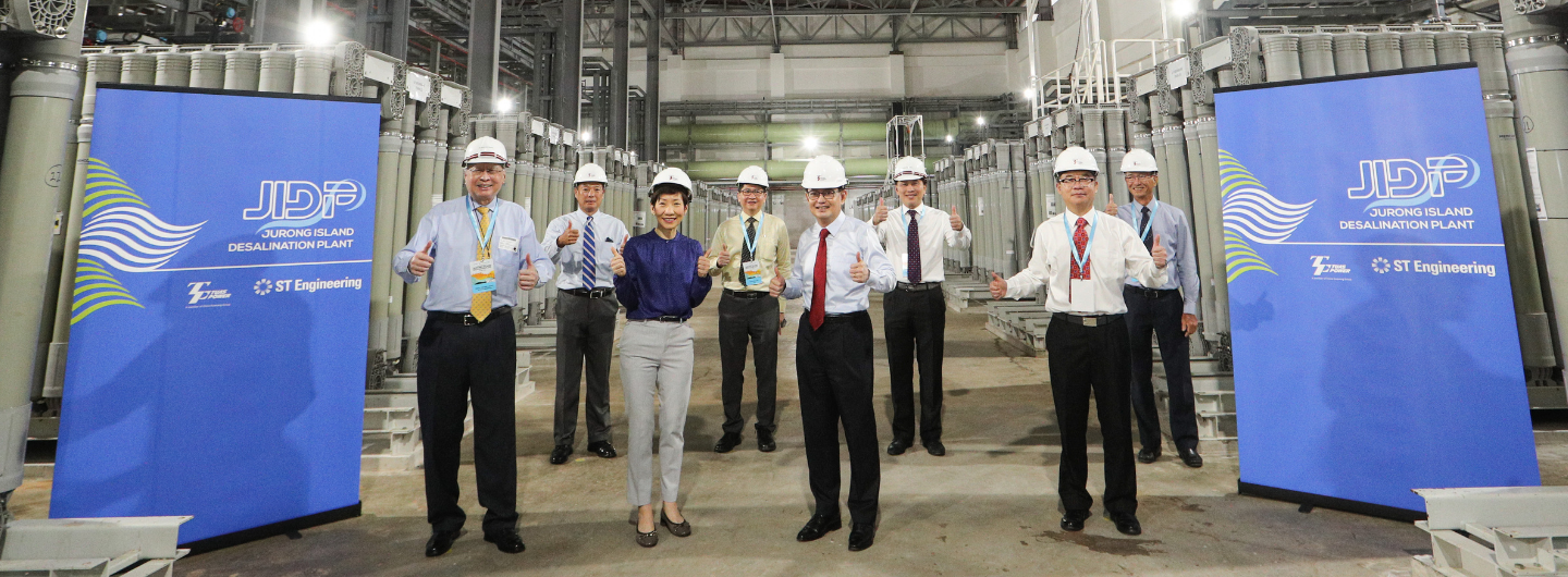 People in hard hats giving thumbs up next to a JIDP/ST Engineering banner.